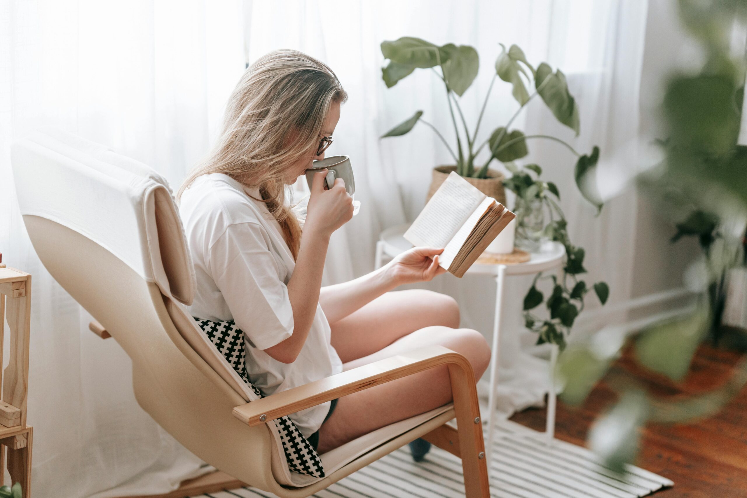 Side view of focused female drinking hot beverage while reading book in armchair in light room with potted green plants